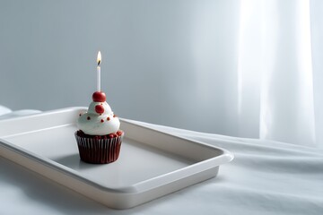 A solitary red velvet cupcake adorned with white frosting and red sprinkles stands on a white tray, illuminated by a single lit candle, creating a minimal and celebratory scene.