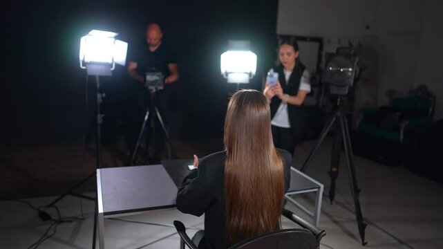 Rear view of a lady with long straight brunette hair sits at desk talking and gesturing in front of camera. Lights of soffits are on the reporter. Cameraman and girl filming on her phone at backdrop.