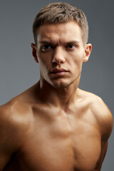 Portrait of a handsome and athletic young man with a defined physique, standing shirtless. He strikes a serious and confident pose against a neutral gray background, showcasing fitness and style.