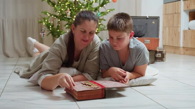 mom and son in casual clothes lying on marble floor opening large vintage book smiling warmly cozy festive indoor setting background featuring christmas tree with lights family moment together