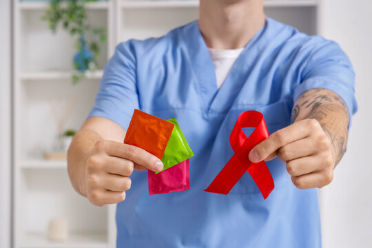 Young doctor showing condoms and red ribbon in clinic, closeup