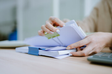 Hands of businessman hold pen for work in stacks of paper files, information search for business reports and heaps of incomplete documents is achieved on a laptop computer table in a modern office