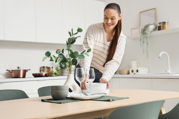 Young woman putting clean dishes on dining table in kitchen