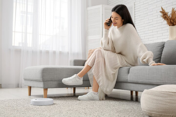 Young woman talking by mobile phone on sofa with robot vacuum cleaner at home