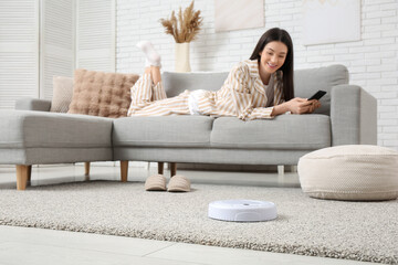 Young woman in pajamas using mobile phone on sofa with robot vacuum cleaner at home