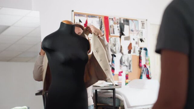 Female designer smiles while sliding beige blazer onto black dress form, chatting with coworker inside studio lined with fabric swatches, sketch filled corkboard, adjusting garment to check fit