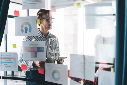 Focused young businessman analyzing charts on glass board in creative office, reviewing data and making strategic notes during planning session