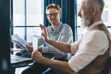 Young businessman laughing while discussing reports with senior colleague in stylish modern office,...
