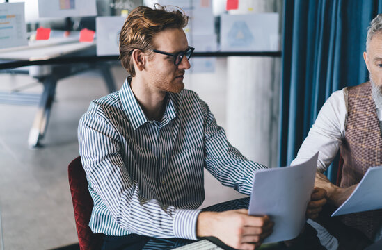 Young businessman attentively reviewing printed reports at glass desk next to senior colleague during team strategy session in open office