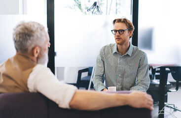Fototapeta na wymiar Young businessman attentively listens to senior mentor during private office conversation, both seated in relaxed atmosphere with natural light and modern interior