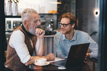 Mature businessman and young male freelancer discussing project at cozy cafe table with laptop and coffee, focused on teamwork and strategy