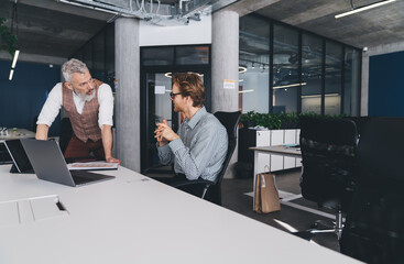 Senior male manager leaning over desk talking to young male colleague during project review in...