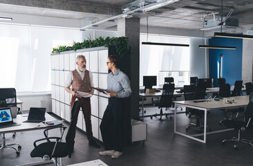 Businessmen exchanging ideas while standing in open plan office, holding printed charts and tablets, discussing project insights in bright space with modern workplace design