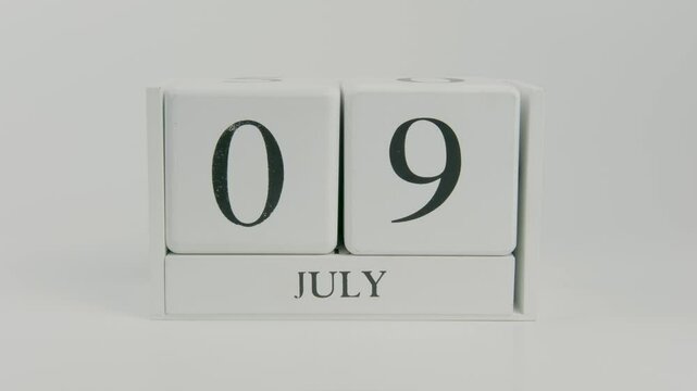 A hand flips a wooden calendar from July 9th to July 10th on a white background, marking the passage of time