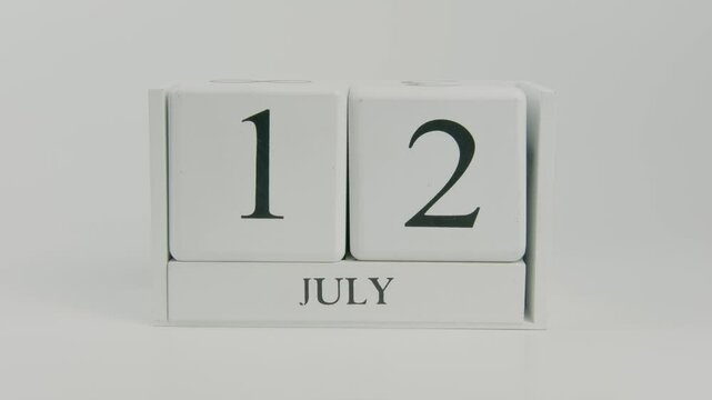 A hand flips a wooden calendar from July 12th to July 13th on a white background, marking the passage of time