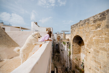 A woman and her big white dog lean over a rooftop ledge, gazing at a historic Italian street below....