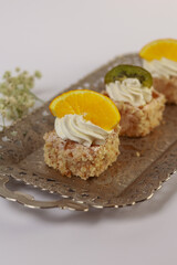 three delicate bite-sized square cake pieces topped with whipped cream and garnished respectively with orange slice, kiwi segment, and another citrus wedge, all artfully arranged on an ornate silver