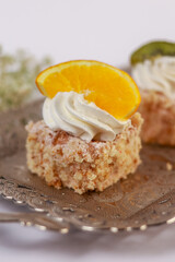 three delicate bite-sized square cake pieces topped with whipped cream and garnished respectively with orange slice, kiwi segment, and another citrus wedge, all artfully arranged on an ornate silver