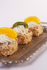 three delicate bite-sized square cake pieces topped with whipped cream and garnished respectively with orange slice, kiwi segment, and another citrus wedge, all artfully arranged on an ornate silver