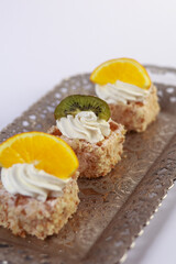 three delicate bite-sized square cake pieces topped with whipped cream and garnished respectively with orange slice, kiwi segment, and another citrus wedge, all artfully arranged on an ornate silver