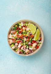 Fresh and vibrant peruvian seafood ceviche with shrimps, octopus, tomatoes, cucumber and parsley, served with lime in a bowl on a light blue background from above, dish of Peru