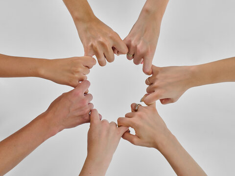 Six hands forming a circle by interlocking their thumbs against a white background. Some hands have rings.