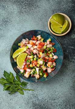 Fresh and vibrant peruvian seafood ceviche with shrimps, octopus, tomatoes, cucumber and parsley, served with lime in a bowl on a stone background from above, dish of Peru