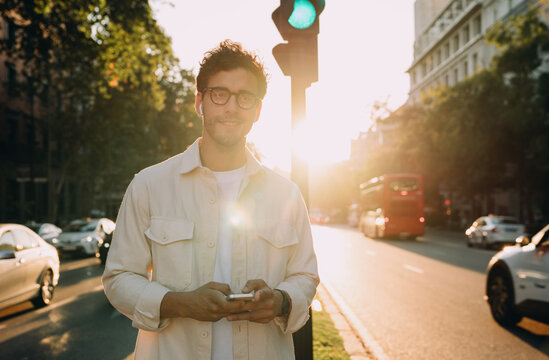 stylish businessman stands on bustling city street at sunset, holding smartphone. His confident expression and smart attire reflect success, modern entrepreneurship, and seamless digital connectivity - Powered by Adobe