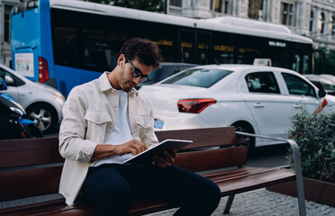 young entrepreneur in stylish attire engages with tablet while sitting on bench near busy street. Focused and determined, he embraces the flexibility of remote work in a fast-paced urban environment