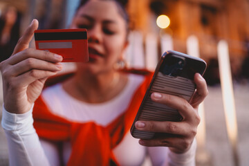 Confident young woman makes a secure online payment with her credit card and smartphone. The vibrant urban setting and warm lights emphasize modern financial freedom and digital shopping convenience