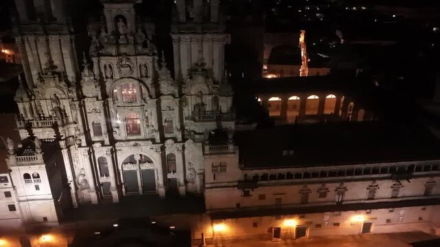 4K drone footage captured at night showing a wider aerial view of the Plaza del Obradoiro and the Cathedral of Santiago de Compostela in Galicia, Spain. The video highlights the illuminated main squar