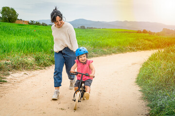 Toddler girl learning to ride a bike outdoors with her mother's help. Concept of baby girl growing and developing, mother daughter bonding together outdoors parenting