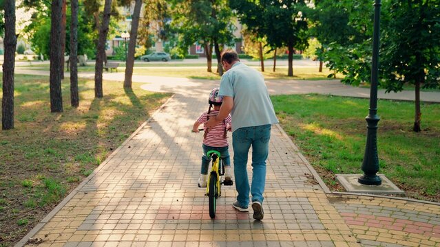 Happy family, little girl in helmet together with her dad learns to ride bicycle outdoors in spring. Child cyclist with Father, nature. Family weekend. Father teaches kid daughter to ride bike in park