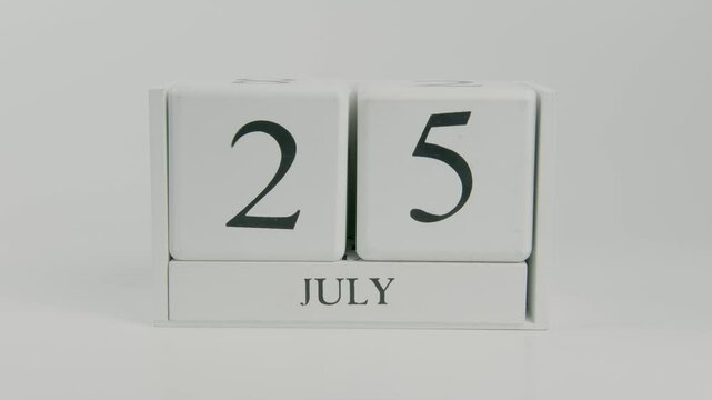 A hand flips a wooden calendar from July 25th to July 26th on a white background, symbolizing the passage of time and the ongoing shift from one day to the next
