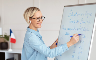 Friendly female teacher writing grammar rules on blackboard during online French lesson. Positive young tutor teaching foreign language on web. Remote education and tutoring concept