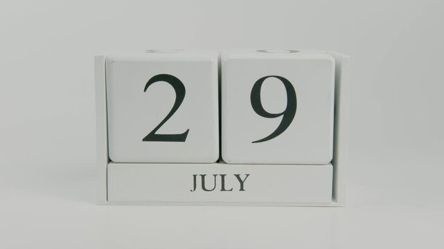 A hand flips a wooden calendar from July 29th to July 30th on a white background, highlighting the passage of time