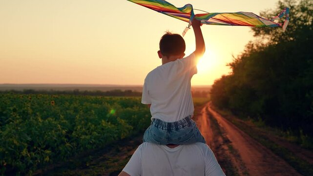 Happy boy on dad shoulders runs along country road, plays with toy kite, sunset. Child, dad is running with kite, sun. Kite floats in child hand. Kid with toy kite. Son plays dreams of fly, traveling