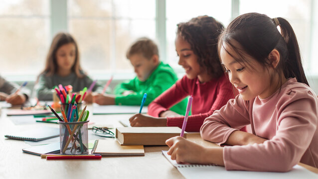 Japanese Schoolgirl Taking Notes Writing Essay Learning In Modern Classroom At School. Multiracial Children Having Class. Knowledge And Education. Selective Focus, Panorama