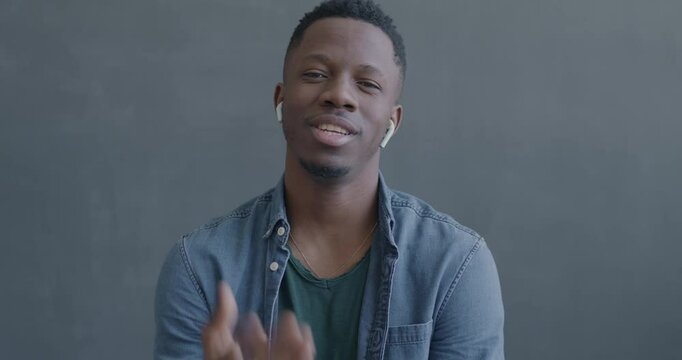 Portrait of African American man making online video call speaking and gesturing looking at camera using wireless earphones on black color background.