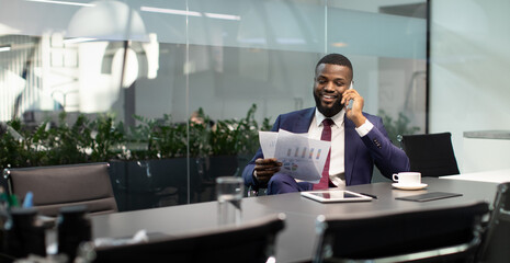 Fototapeta na wymiar Wealthy black guy in suit entrepreneur having phone conversation with his assistant or business partner, sitting at table with digital tablet on, looking at documents, panorama with copy space