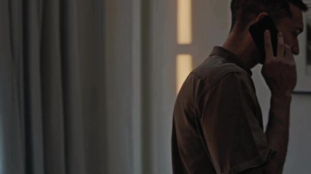 Stressed man talking on a smartphone near a window in a dark room. Anxiety, difficult conversation, bad news, loneliness and emotional pressure concept.