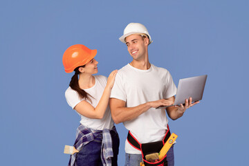 Cheerful young couple in protective hardhats using laptop computer while posing on blue studio background, checking renovation project for their apartment, searching repair tools online in internet