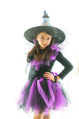 Cute girl in a witch costume and glasses poses standing in front of a large bright panoramic window. Concept of a children's party and childhood