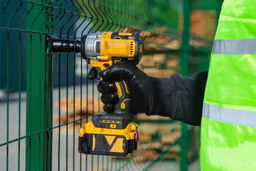 Worker in a safety vest holds a impact wrench near a green construction site fence. Industrial labor and safety gear.