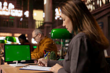 Mature female student reading and writing at desk next to green screen, studying documents and doing her research to use important citations in her master degree thesis. Bookworm.