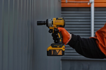 A worker in a protective vest and gloves presses an impact wrench against a metal wall, demonstrating the workflow on a construction site.