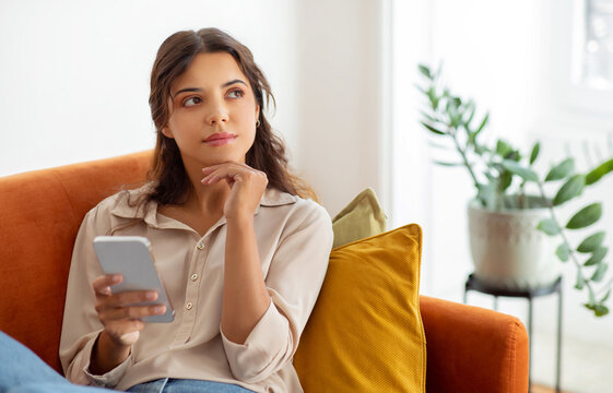 Pensive young woman holding smartphone and looking away while relaxing on couch at home, beautiful female deep in thoughts using mobile phone in domestic interior, thinking and planning something