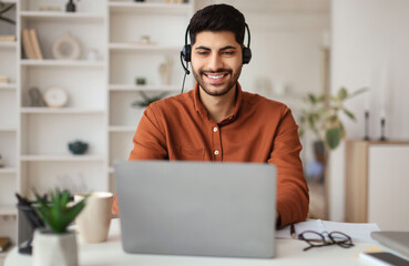 Portrait Of Smiling Middle Eastern Male Manager In Headset Working On Laptop Computer In Modern Home Office, Happy Guy Sitting At Desk And Consulting Clients Online Using Pc, Free Copy Space