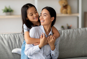 Happy asian little girl hugging her beautiful mother and smiling to each other, sitting on sofa in...