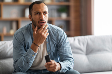 Shocked Black Man With Remote Controller In Hand Sitting On Couch At Home, Confused Young African...